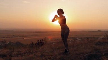 Genç bir kadın gün batımında dışarıda çömeliyor, sağlıklı bir yaşam tarzı ve spor hobisi.
