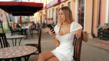 Güzel sarışın bir kadının yakın plan portresi. Çekici gözleri var. Elinde akıllı bir telefon, bir sokak kafesinde bir masada oturuyor ve tatlı tatlı gülümsüyor.
