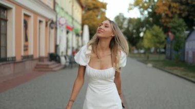 Çeşitli alışveriş çantalarıyla beyaz bir yaz elbisesi içinde muhteşem bir genç kadın gün batımında caddede yürüyor. Yavaş çekim.
