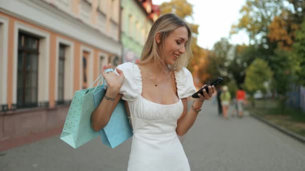 快乐的年轻购物狂女人拿着纸袋购物 走在城市街道上 快乐的女士在黑色星期五用智能手机购物 — 图库视频影像