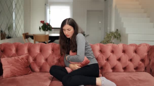 月経痛に苦しむ病気の若い女性 痛みを伴う胃の痛みやソファに座っている期間のけいれんを持つ手を圧迫腹を持つ女性 腹部の痛み 痛みのある期間の概念 — ストック動画