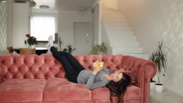 快乐漂亮的年轻女人在舒适的客厅里用智能手机在家里 一个女人躺在舒适的沙发上 她浏览互联网 观看社交媒体上的视频 并进行娱乐活动 — 图库视频影像