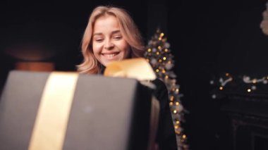 Noel ağacının arka planında altın kurdeleli büyük bir hediye kutusunu tutan genç, neşeli bir kadının portresi. Bir kız Noel hediyesini kabul eder, yakın plan.