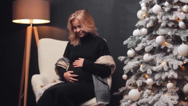 妊娠した若い魅力的な女性は 夕方自宅のクリスマスツリーの近くの椅子に座って 彼女の腹を笑い 楽しい 幸せな時間を持って 不思議に — ストック動画