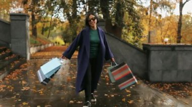 Alışveriş çantalı ve güneş gözlüklü bir iş kadını, mavi bir ceket giyiyor, sonbahar parkında arka planda geziyor, indirimler ve satış sezonunda şık bir iş kadını.