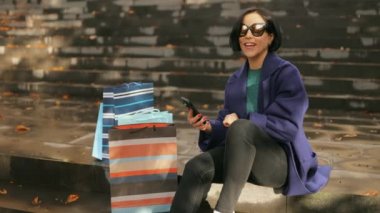 Heyecanlı orta yaşlı kadın alışverişçisi, elinde alışveriş çantalarıyla geziyor, cep telefonu uygulamasını kullanıyor, nakit para alıyor, e-ticaret mağazasından moda kıyafetleri alıyor.