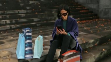 Şık, orta yaşlı, kısa saçlı esmer kadın güneşli bir sonbahar gününde alışveriş torbalarıyla parktaki merdivenlerde oturur, akıllı telefon kullanır ve kameraya gülümser.