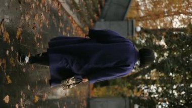 Dikey video, uzun ceketli, siyah saçlı ve elinde çanta olan çekici orta yaşlı bir iş kadını bulutlu bir günde bir sonbahar parkında yürüyor, gülümsüyor, sonbaharda yürüyüş yapıyor.
