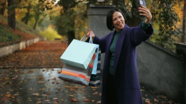 一位美丽 快乐的中年妇女提着购物袋走在城市公园的中央 在一个手机视频电话上谈话 并展示她的购买情况 — 图库视频影像