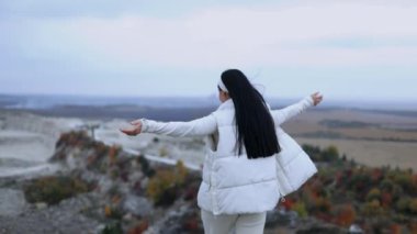 Uzun saçlı genç bir kadın bir tepenin üstünde duruyor ve kollarını yanlara kaldırıyor. Turist bir kız sonbahar doğasının tadını çıkarıyor, dağlarda yalnız bir gün.
