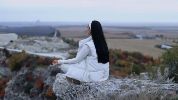崖の端に座ったヨガの位置に座っている若い女性 山の上に リラックスして眺めている 秋の自然の中を旅しながらリラックスした瞑想をする女性 — ストック動画