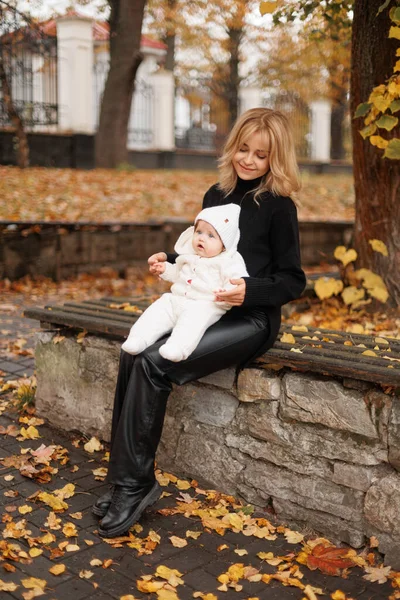 Genç, gülümseyen bir anne, küçük kızını kollarına alır. Sonbahar doğasında, şehir parkında, bir portre.
