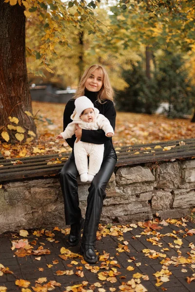 Güneşli bir günde, sonbahar parkında kollarında genç ve güzel bir anne ve kızının portresi, kameraya yakından bakıyor.