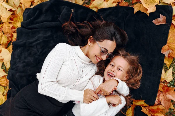 Genç anne ve kız sonbahar yaprakları üzerinde parktaki battaniyeye uzanıyor, sarılıyor ve gülümsüyor, dışarıda eğleniyorlar.