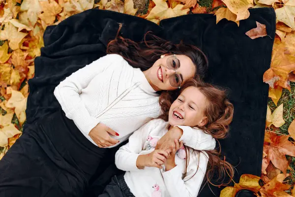 Genç anne ve kız sonbahar yaprakları üzerinde parktaki battaniyeye uzanmış kameraya bakıyor ve gülümsüyor, dışarıda eğleniyorlar.