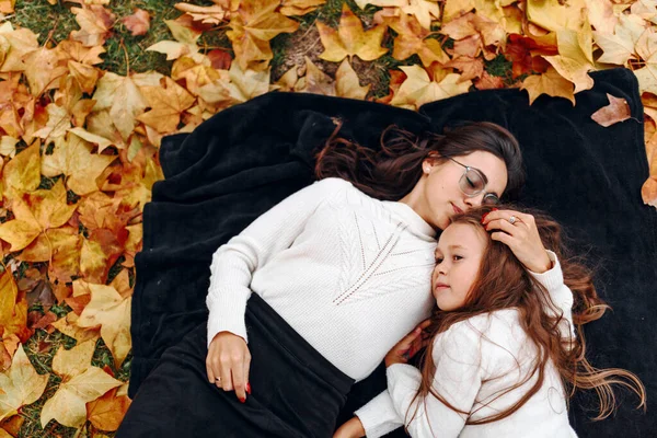 Genç anne ve kızı sonbahar yaprakları üzerinde parkta bir battaniyeye uzanmış, sarılıyor ve gülümsüyor, dışarıda eğleniyorlar, anne ve kız birbirlerini seviyorlar.