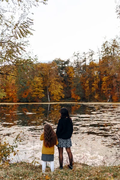 Anne ve küçük kızı sonbahar parkında gölün kenarında duruyorlar. Eğlence yürüyüşü, anne ve kızının sevgisi.