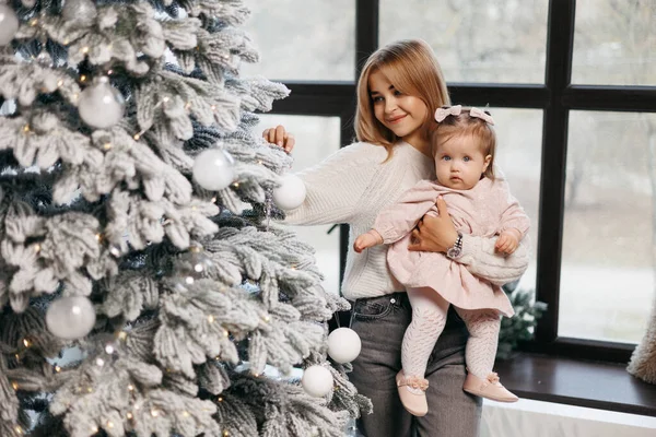 mother and little daughter stand near the Christmas tree and decorate it. The family celebrates the new year. A mother holds her baby daughter in her arms