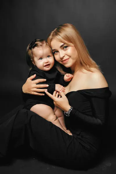 Küçük kızını kollarında tutan genç bir annenin portresi. Stüdyoda siyah bir arka planda sarılıp gülümsüyor. Bir annenin çocuğuna olan sevgisi. Bebek ve anne kameraya gülümsüyor.