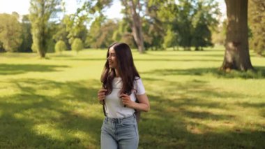 Young woman girl tourist student with a backpack walks in the park outdoors on a summer day, admires nature, looks around and smiles happily at the camera