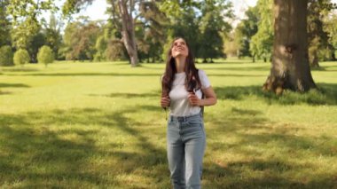 Young woman girl tourist with a backpack walks in a park or grove outdoors on a summer day, admires nature, looks up and blocks the sun's days with her hand