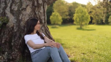 Young attractive woman girl sitting in the grass leaning on a big tree and enjoying nature on a warm sunny summer day, tourist traveler, student, vacation, concept of freedom and love of nature