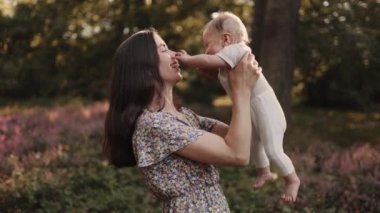 Genç, çekici bir anne ve mutlu bir erkek çocuk ormanda kır çiçekleriyle oynuyor ya da gün batımında park ediyor.