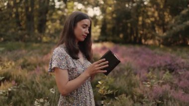 Genç Hıristiyan bir kadın, dışarıdaki bir parktaki kır çiçeklerinin çayırında İncil kitabı okuyor ve Tanrı 'ya şükranla cennete bakıyor.