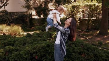 Mutlu ve duygusal genç bir anne sevimli bebeğini kollarına alır ve onu fırlatır. Birlikte dışarıda vakit geçirmek, güneşli bir gün geçirmek ebeveynlerin ve çocukların mutlu anneleri olmak.