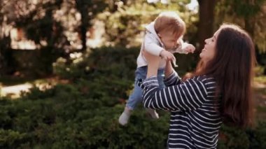 Sonbaharda güneşli bir parkta yürüyüş yapan küçük bir çocuğu olan genç bir anne. Anne oğluna sarılıyor ve onunla dönüyor. Mutlu çocukluk ve annelik aşkı kavramı, yavaş çekim, arka planda güzel doğa.