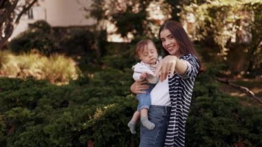 Genç ve sevimli bir anne kucağında bebekle çocuğun dikkatini çekmek için kamerayı işaret ediyor ve parkta el sallıyor.