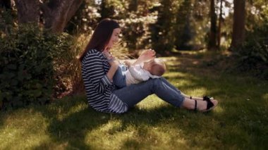 Genç anne parkta çimenlerde oturur ve küçük oğluyla oynar, bacaklarını öper, gülümser ve sarılır. Mutlu annelik. Dinlen, çocukla vakit geçir.