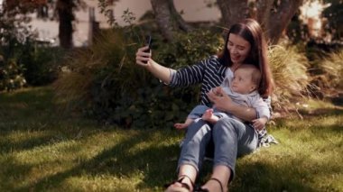 Gülümseyen anne akıllı telefonuyla güzel bir video görüşmesi yapıyor. Küçük oğluyla parkta çimlerin üzerinde oturuyor. Bebeğiyle yürüyor, kameraya el sallıyor, blog çekiyor, anne blogcu.
