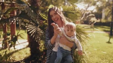 Neşeli ve tatlı bir anne küçük oğlunu kollarında tutuyor, sarılıyor ve birlikte eğleniyor. Mutlu çocukluk ve annelik kavramı. Gülmek, gülümsemek, duygular, hafta sonunun tadını çıkarmak, kameraya bakmak ve el sallamak.