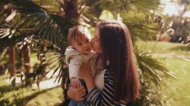 Neşeli ve genç bir anne küçük oğlunu kollarında tutuyor, sarılıyor, öpüşüyor ve birlikte eğleniyor. Mutlu çocukluk ve annelik kavramı. Gülmek, gülümsemek, duygular, hafta sonunun tadını çıkarmak, parkta güneşli bir gün.