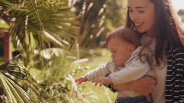 Genç bir anne parkta kollarında bir çocukla yürür, bitkileri ve çalıları gösterir, aile yürüyüşleri ve aktif eğlence kavramı.