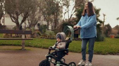 Bebek arabası taşıyan bir annenin yakın çekimi, sonbahar parkında bir bebekle üç tekerlekli bisiklet. Yan görüş. Annelik, temiz havada yürümek, çocuk bakımı. Oğlu etrafındaki dünyayı inceliyor.