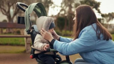 Küçük çocuk parkta üç tekerlekli bebek arabasında oturuyor, annesi yaklaşıyor, elbiselerini tamir ediyor, sarılıyor ve onu öpüyor. Mutlu annelik ve çocuk bakımı