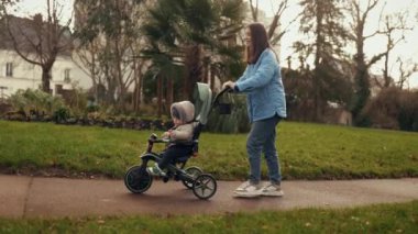 Mutlu gülümseyen anne oğlunu sonbahar parkında trocheoli bisikletine bindiriyor. Yan görüş. Bebekle bir yürüyüş.