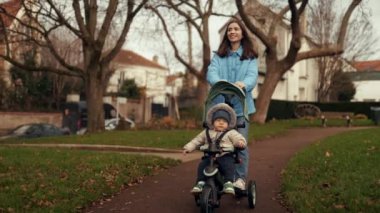Çekici genç anne, yakışıklı oğluyla birlikte sonbahar ya da kış parklarında bir bebek arabasında bisiklet sürüyor. Çocukluk kavramı, mutlu aile ve yaşam tarzı. Doğa, temiz hava, hafta içi aktif eğlence.
