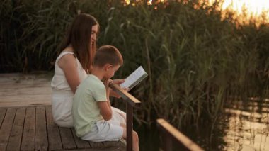 Anne ve oğlu yazın gün batımında bir gölün kıyısında kitap okuyorlar. Anne ve çocuğun doğadaki güzel eğlencesi.