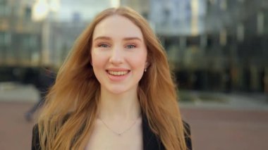 Uzun saçlı ve mavi gözlü şık bir genç kadının yakın çekimi, açık havada güneş ışığında duruyor, yumuşak, kendinden emin bir gülümsemeyle modern ve profesyonel şehir portresiyle kameraya bakıyor..