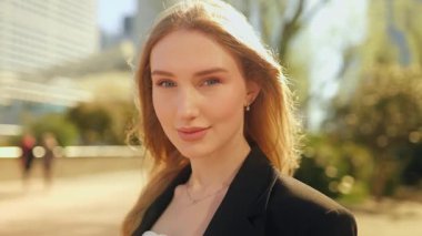 Uzun saçlı ve mavi gözlü genç bir iş kadınının yakın plan portresi, kameraya sıcak bir şekilde gülümserken parlak güneş ışığı altında kendinden emin ve ışıl ışıl kentsel görünümlü.