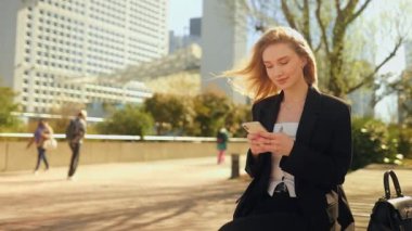 Genç bir kadın bankta oturuyor, akıllı telefonuna odaklanıyor, sonra kameraya samimi ve samimi bir şekilde bakıyor ve gülümsüyor..