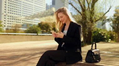 Kendine güvenen, uzun saçlı ve mavi gözlü bir iş kadınının orta ölçekli yakın çekimi, güneşli bir şehir bankında otururken akıllı telefonuna nazikçe gülümseyerek kentsel bir ortamda dijital temasta bulunuyor..