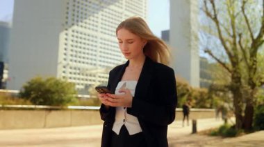 İş elbisesi içinde uzun dalgalı saçlı genç bir kadın elinde akıllı bir telefonla sokakta yürüyor, ekrana bakıyor ve yazı yazıyor ya da interneti kullanıyor.