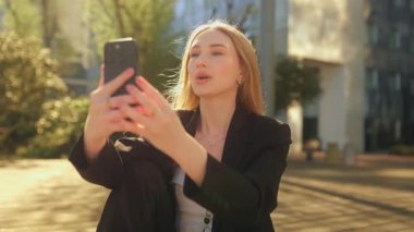 Mavi gözlü, uzun saçlı, gülümseyen genç bir iş kadınının yakın çekimi. Video görüşmesi sırasında bankta oturuyor. Şehir profesyonel ortamında neşeli, doğal bir şekilde gülüyor..