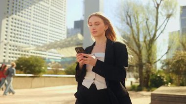 Takım elbiseli, uzun saçlı, kendine güvenen genç bir kadın elinde akıllı bir telefonla sokakta yürüyor, ekrana bakıyor ve yazı yazıyor ya da interneti kullanıyor.