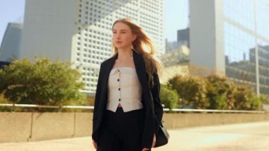 Uzun saçlı, mavi gözlü, resmi giysiler içinde Paris 'te modern bir iş bölgesinde yürüyen, özgüvenli genç bir iş kadını, odaklanmış bir ifadeyle ileriye bakıyor kurumsal başarı ve şehir zarafeti kavramı..