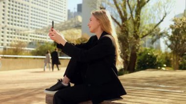 İş kıyafetleri içinde neşeli genç bir kadın şehir bankında oturuyor, telefonuyla bir video görüşmesi yapıyor, gülüyor ve sohbetin tadını çıkarıyor şehir ortamında kurumsal yaşam tarzını rahatlatıyor..